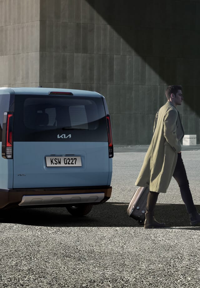 Dreiviertel-Rückansicht des Kia PV5 Passenger, geparkt in der Nähe eines Gebäudes, während ein Mann vorbeigeht.